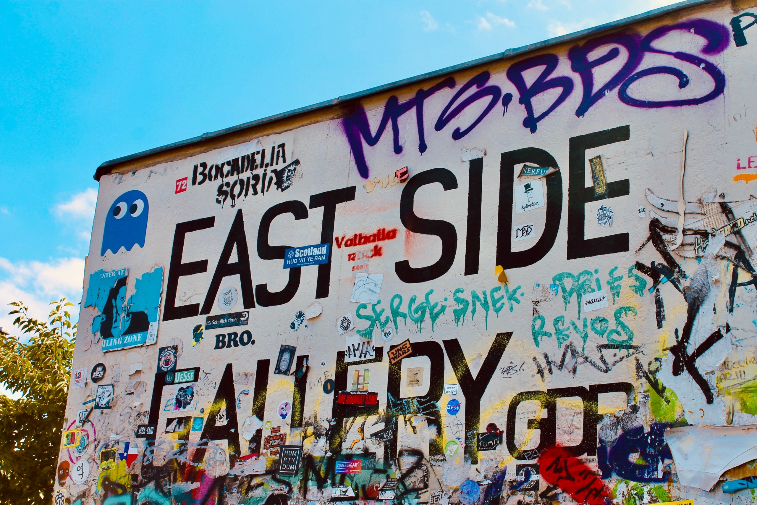 © Marie Rouilly Graffiti-covered wall with bold "EAST SIDE GALLERY" text, colorful tags, stickers, and artwork, conveying urban creativity and vibrancy under a blue sky.