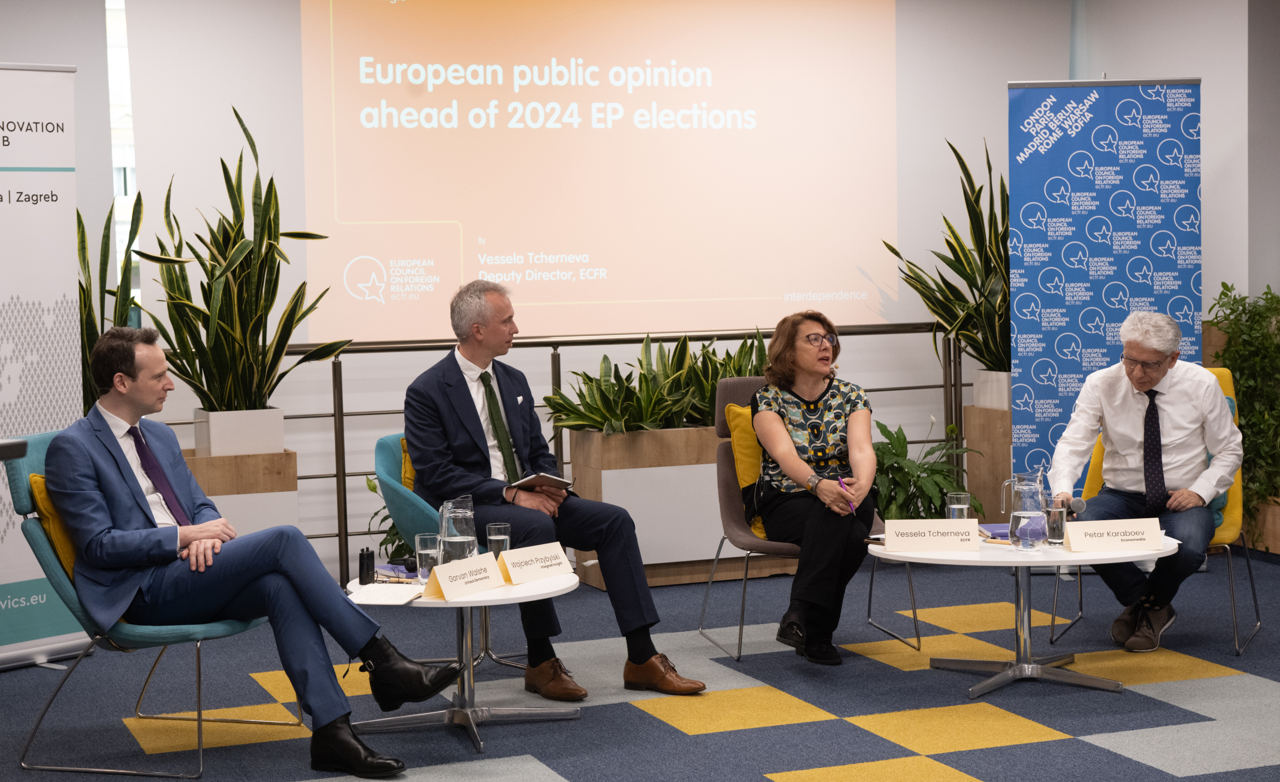 Four people are seated in a discussion panel on European public opinion ahead of 2024 elections. They sit on blue and yellow chairs with plants and banners in the background.