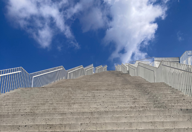 DISCOVER Concrete stairs lead upward to a bright blue sky with fluffy clouds. White railings line the stairs, creating a sense of openness and aspiration.