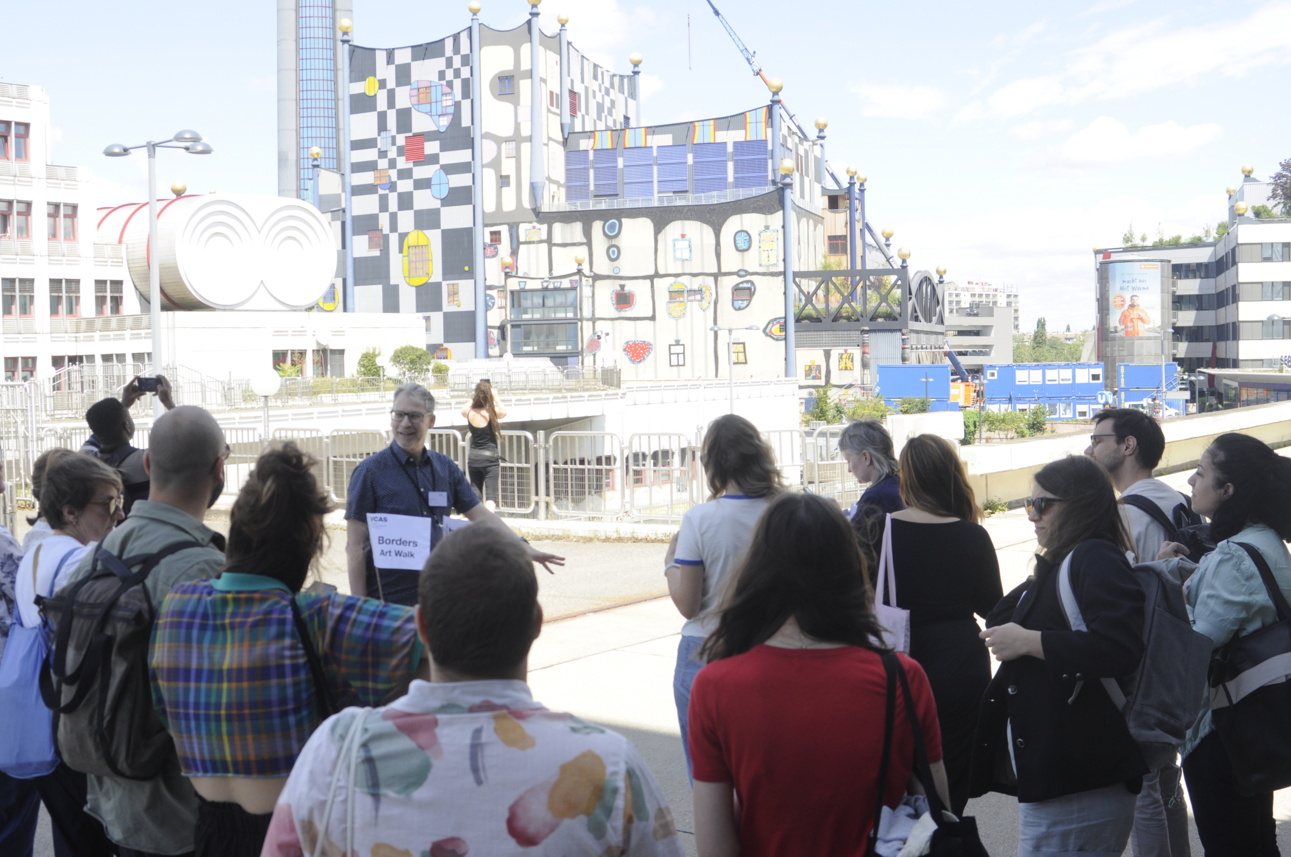 A group of people on an art walk stand before a vibrantly painted building featuring colorful geometric and abstract patterns, under a clear sky.