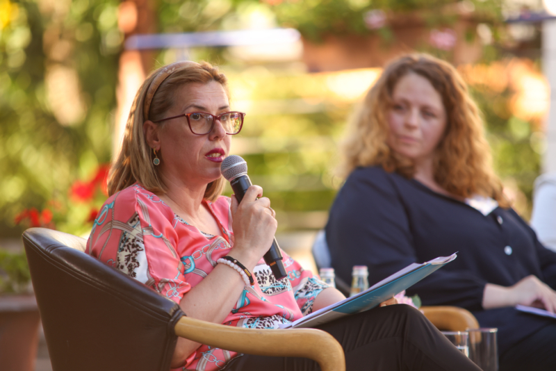 A lady speaking in a garden holding a microphone and speaking at the Exploring "BORDERS": A preview of the NECE Festival 2024 in Tirana