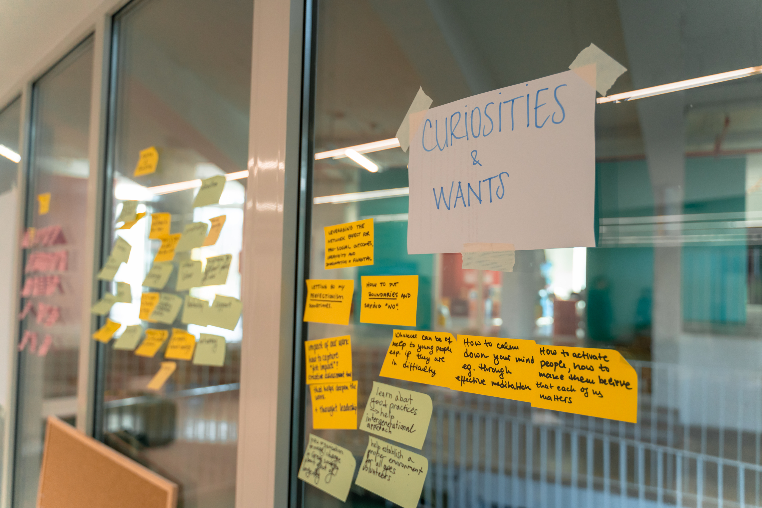 © THE CIVICS Innovation Hub | Geri Lula Glass wall covered in yellow and blue sticky notes with handwritten ideas under a paper labeled “Curiosities & Wants,” creating a brainstorming atmosphere.