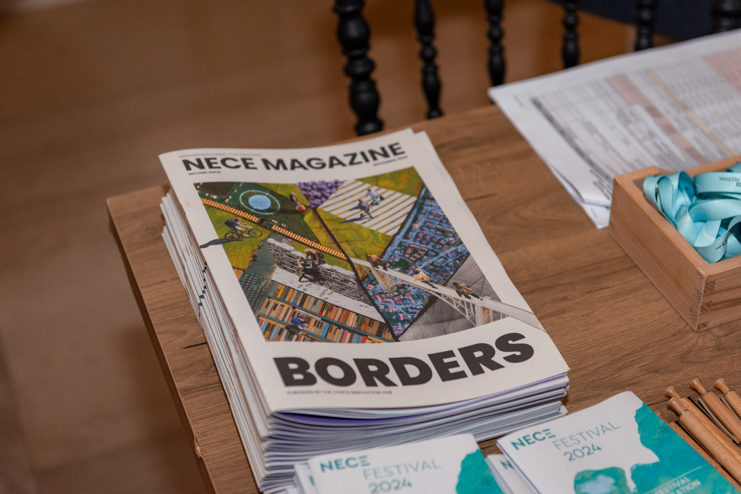 © THE CIVICS Innovation Hub | Geri Lula Stack of NECE Magazine issues titled "BORDERS" on a wooden table. Nearby are NECE Festival 2024 brochures, pens, and a box of teal ribbons.
