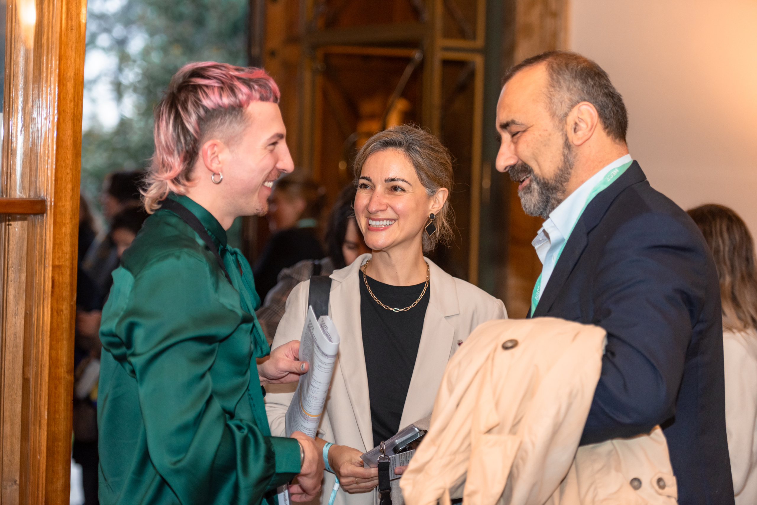© THE CIVICS Innovation Hub | Geri Lula Three people are warmly conversing indoors at NECE Festival 2024. The person on the left has pink hair and wears a green shirt. The mood is friendly and engaged.