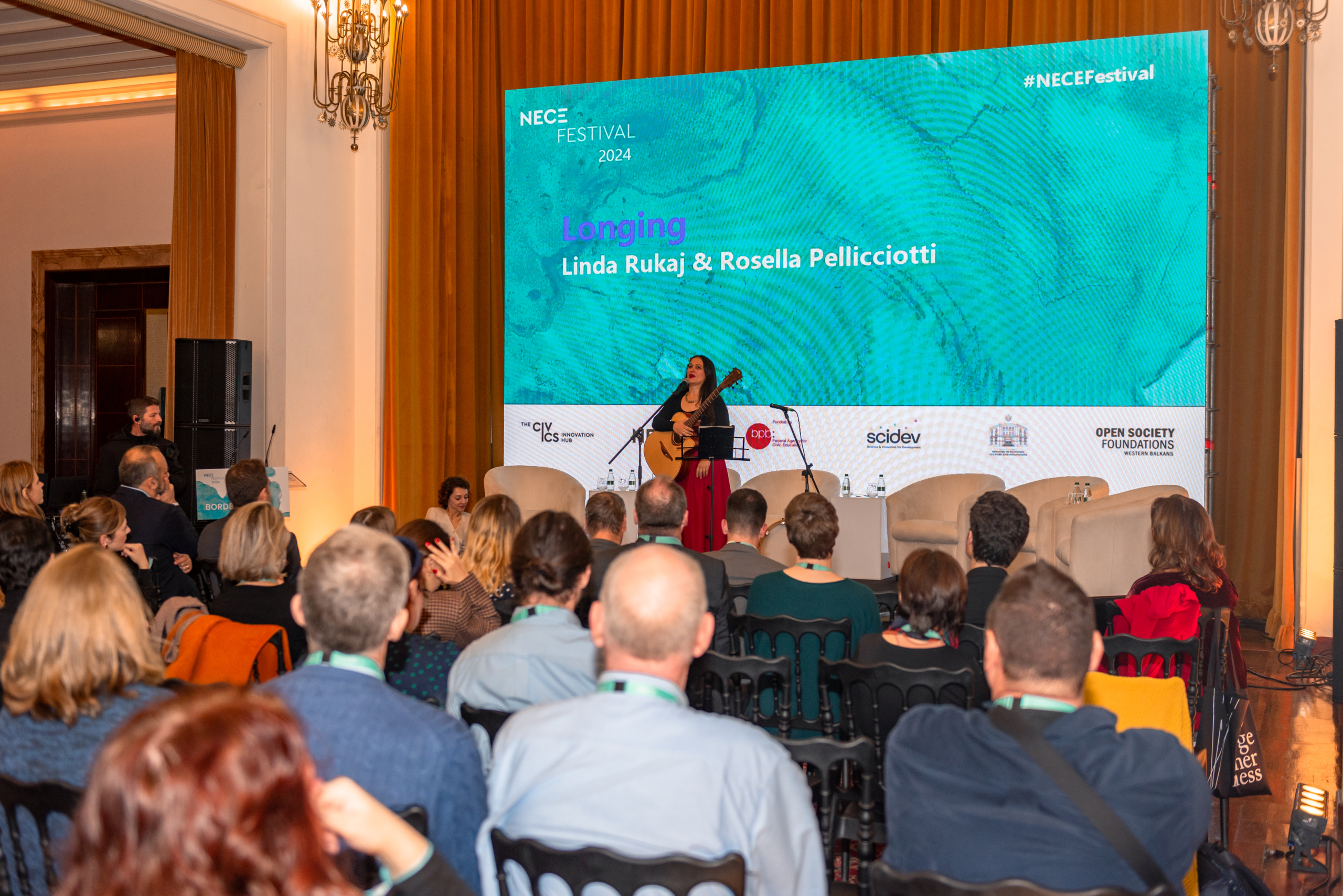 © THE CIVICS Innovation Hub | Geri Lula Musician on stage performing with a guitar at the NECE Festival 2024. Audience seated, large screen behind displays event details.