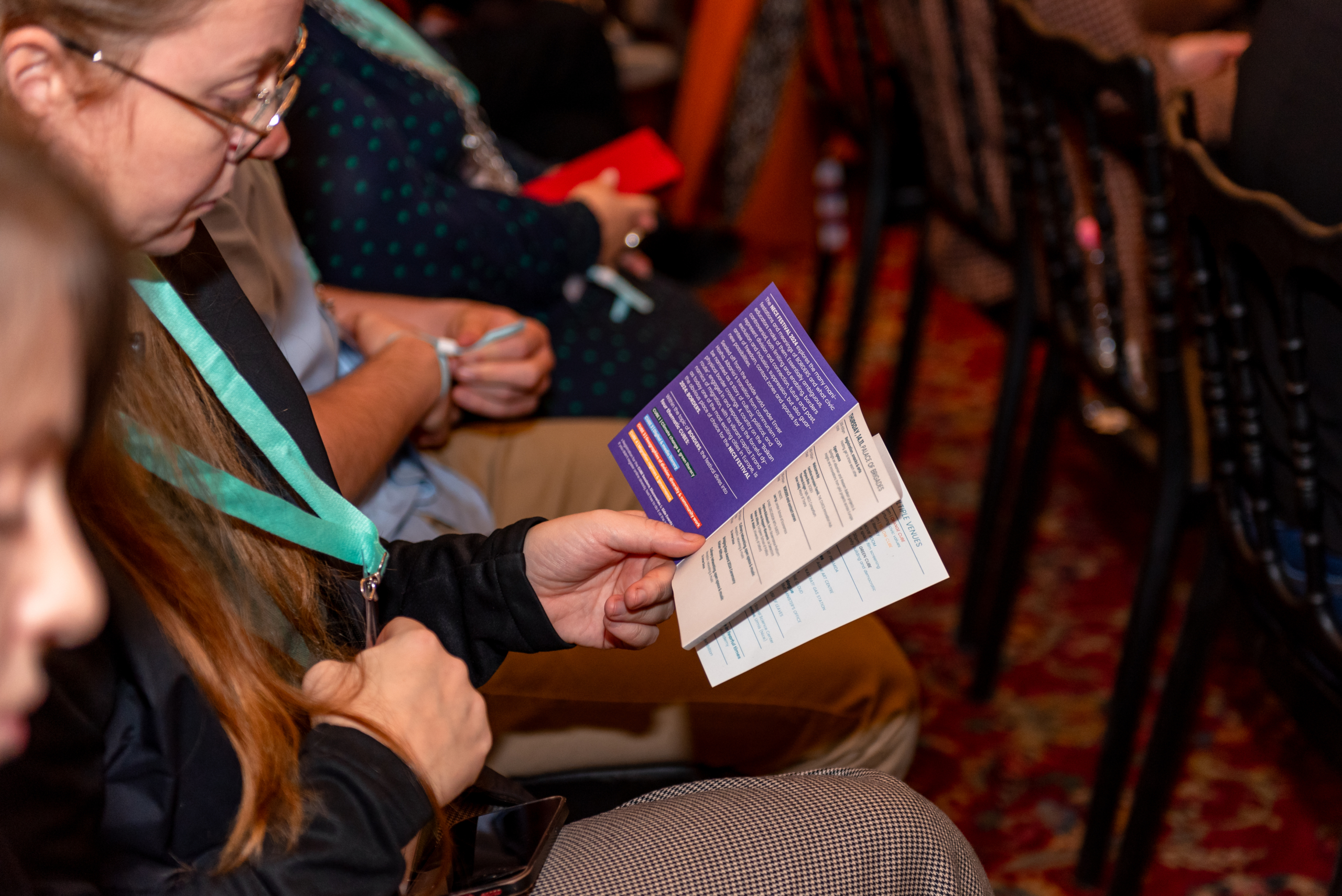 © THE CIVICS Innovation Hub | Geri Lula A person in glasses holds a purple pamphlet, sitting among an attentive audience at NECE Festival 2024.