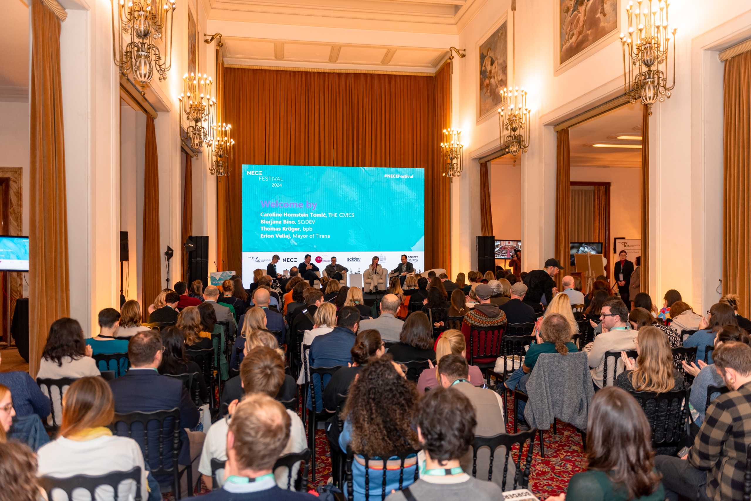 © THE CIVICS Innovation Hub | Geri Lula A large conference room filled with seated attendees facing a stage at NECE Festival 2024. A screen above the panelists displays names and welcomes the audience.