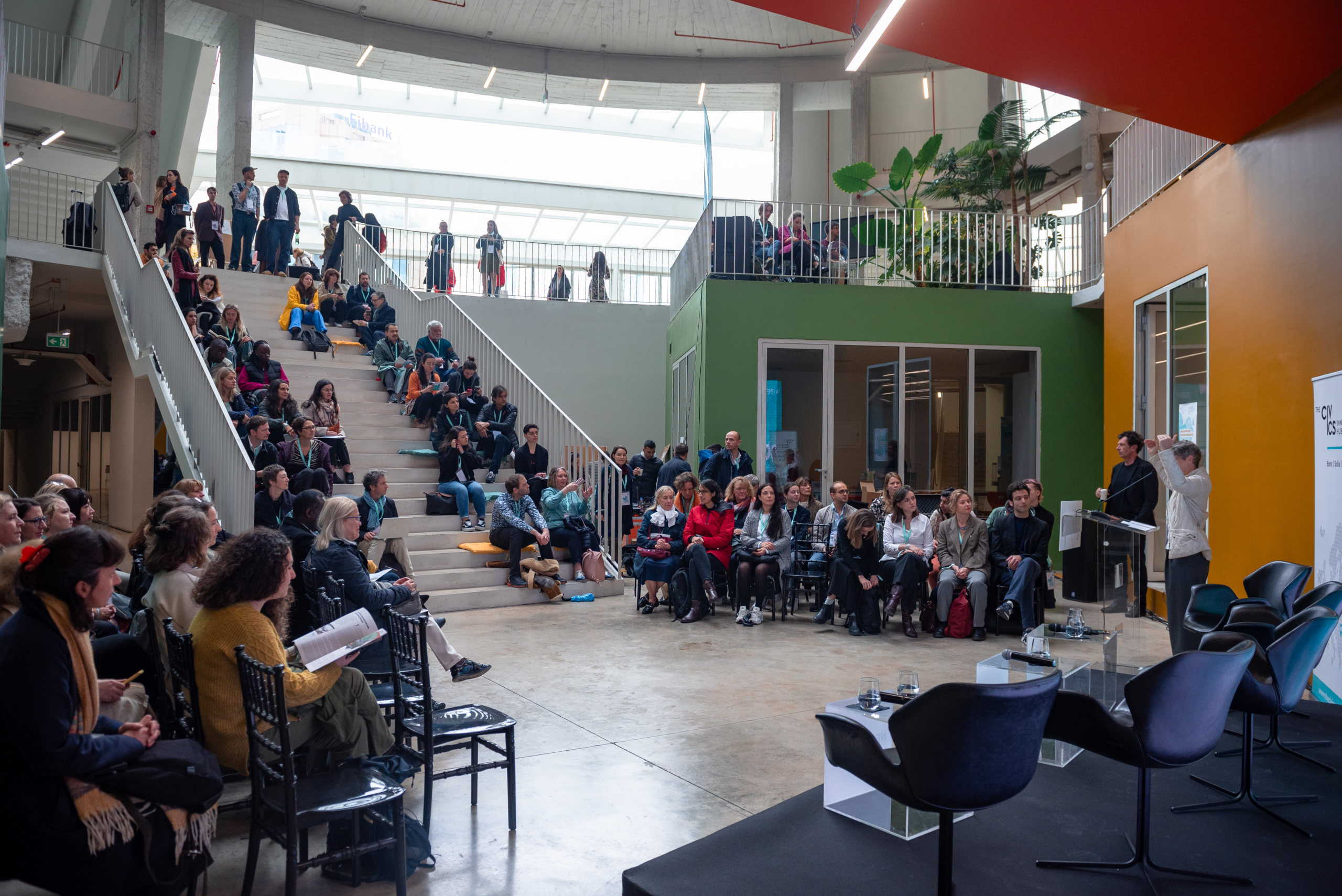 © THE CIVICS Innovation Hub | Geri Lula A diverse audience attentively listens in a modern atrium, seated on stairs and chairs during NECE Festival 2024.