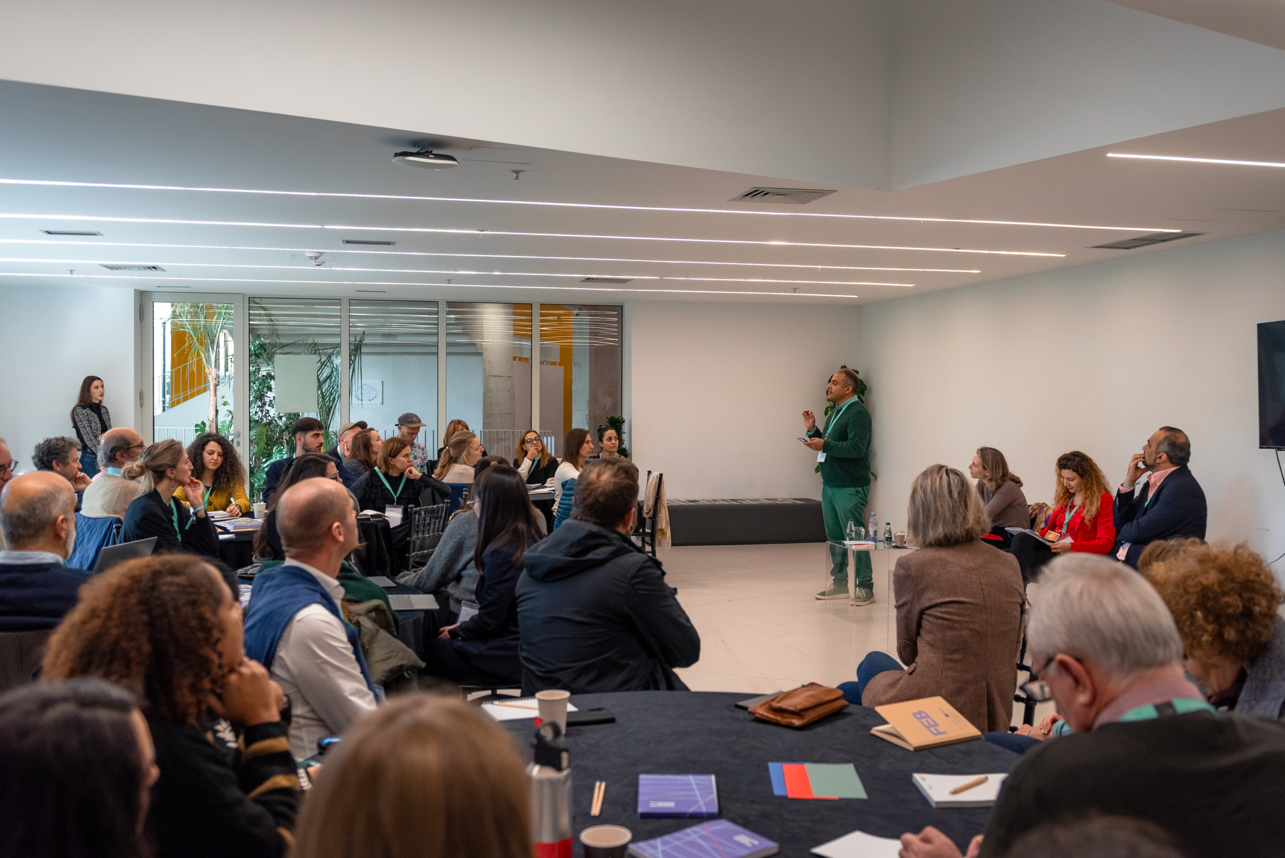 © THE CIVICS Innovation Hub | Geri Lula A presenter in green speaks to a seated audience in a modern conference room during NECE Festival 2024. Attendees appear engaged, some seated at tables with notebooks.