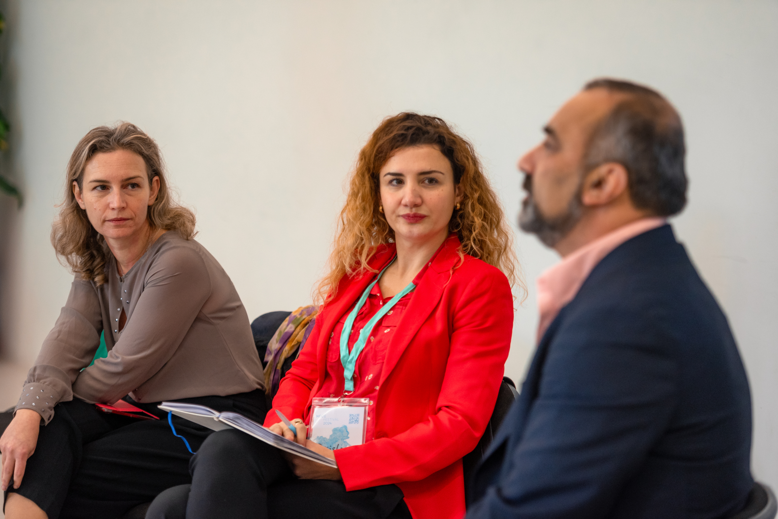 © THE CIVICS Innovation Hub | Geri Lula Three people are engaged in a discussion. A woman in a red blazer holds a notepad, looking attentive, while another listens intently.