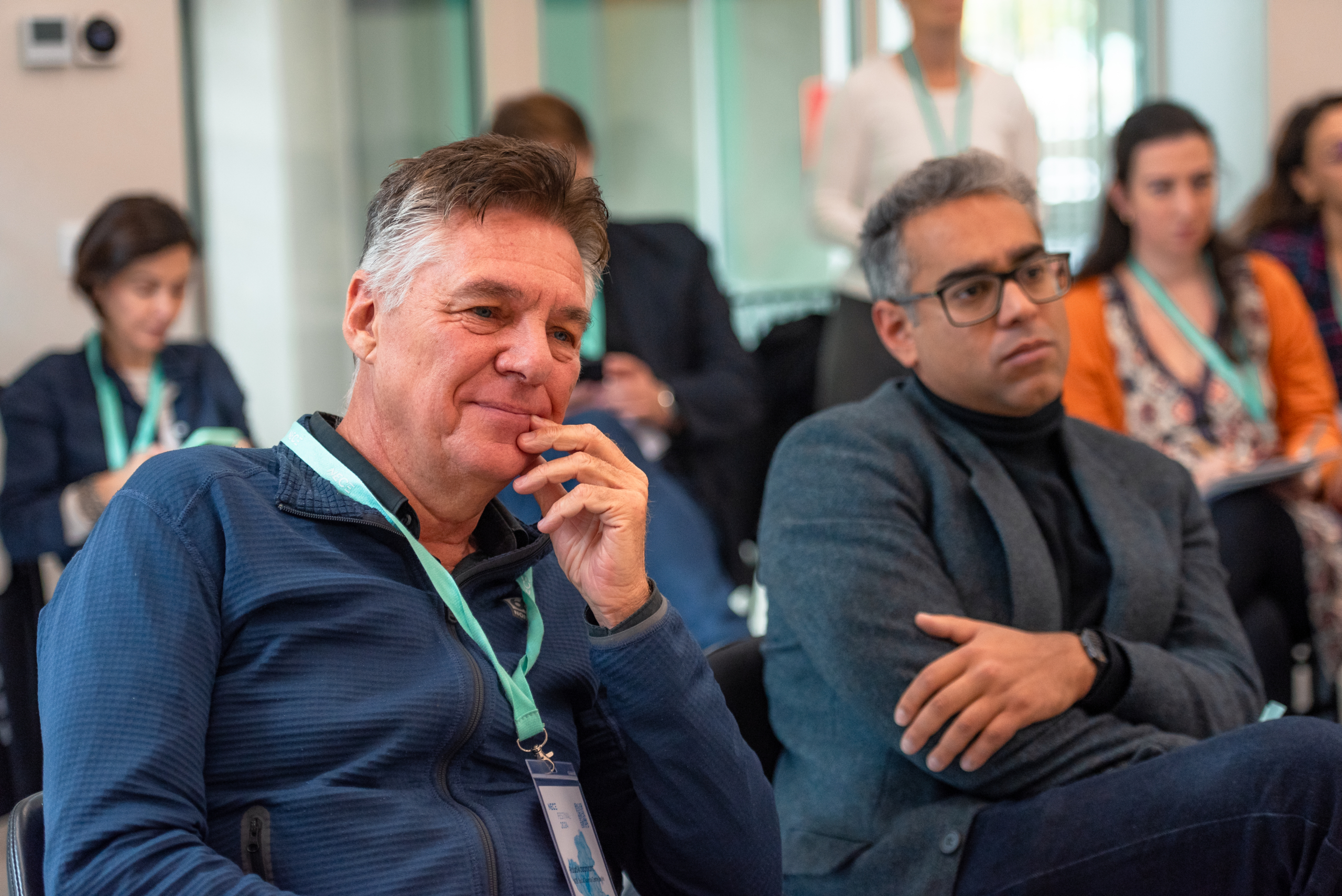© THE CIVICS Innovation Hub | Geri Lula Two men sit attentively in a classroom setting during NECE Festival 2024, both wearing lanyards. The atmosphere is focused, with others in the background listening intently.