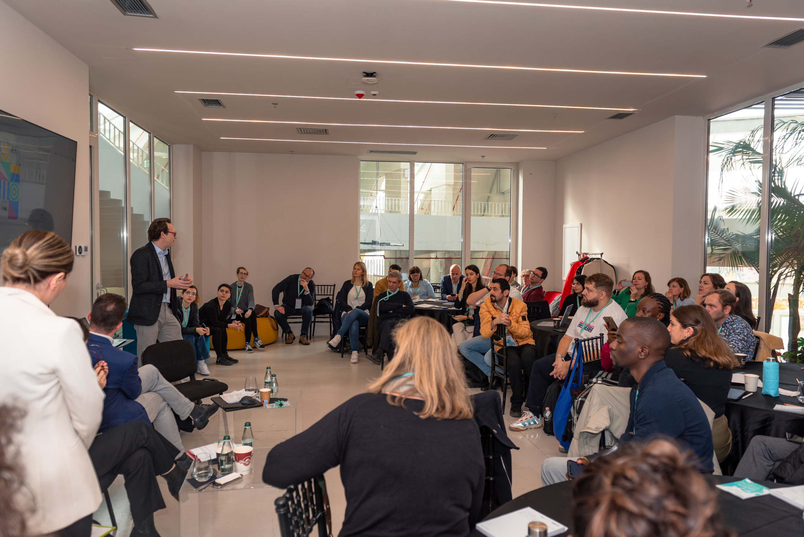 © THE CIVICS Innovation Hub | Geri Lula A man speaks in front of a diverse group sitting in a well-lit room, engaging in a collaborative workshop during NECE Festival 2024.
