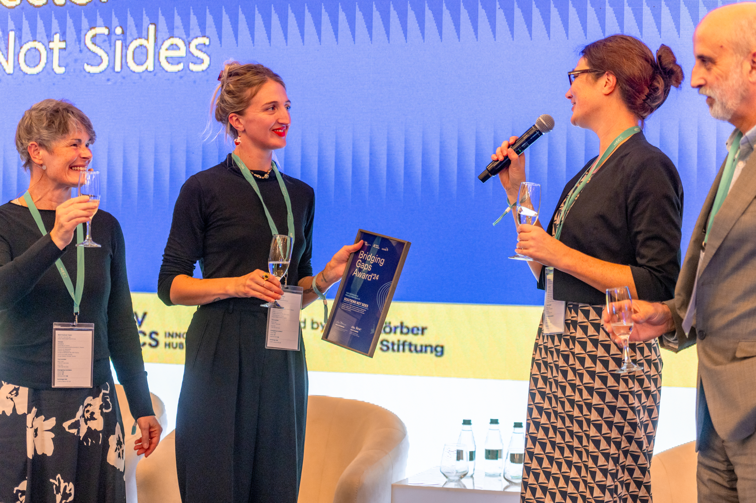 © THE CIVICS Innovation Hub | Geri Lula A group of four people on a stage, holding champagne glasses and smiling. One person holds a microphone, while another holds an award plaque.