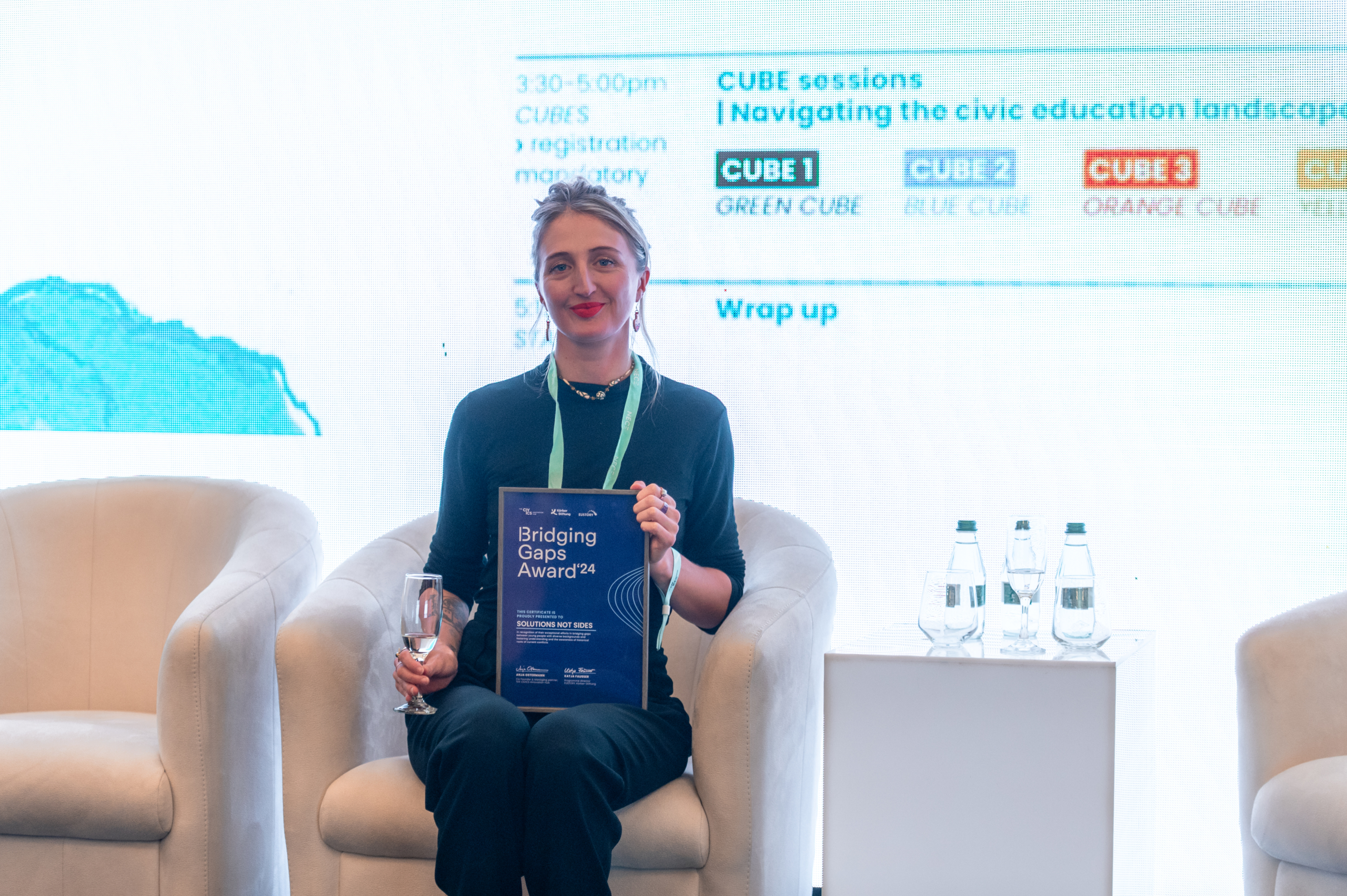 © THE CIVICS Innovation Hub | Geri Lula A person sits between white armchairs, smiling and holding a "Bridging Gaps Award '24."