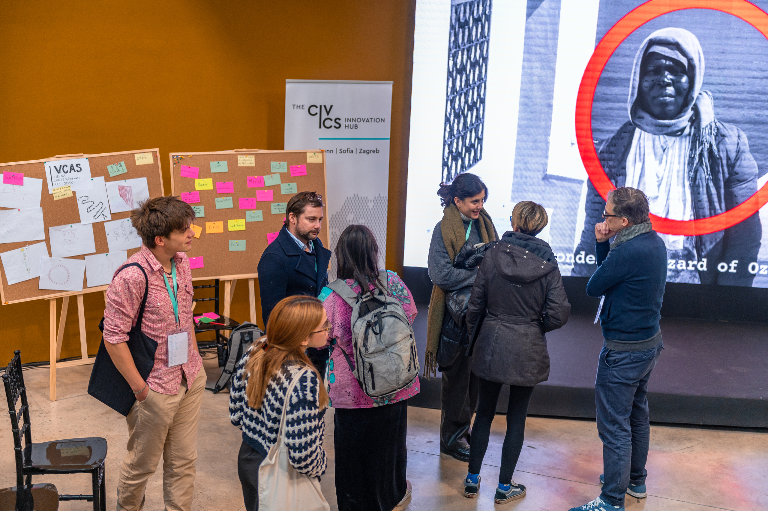© THE CIVICS Innovation Hub | Geri Lula A group of people engage in conversation at NECE Festival 2024. Behind them, corkboards display colorful sticky notes and drawings. A large screen shows a black-and-white photo with a red circle.