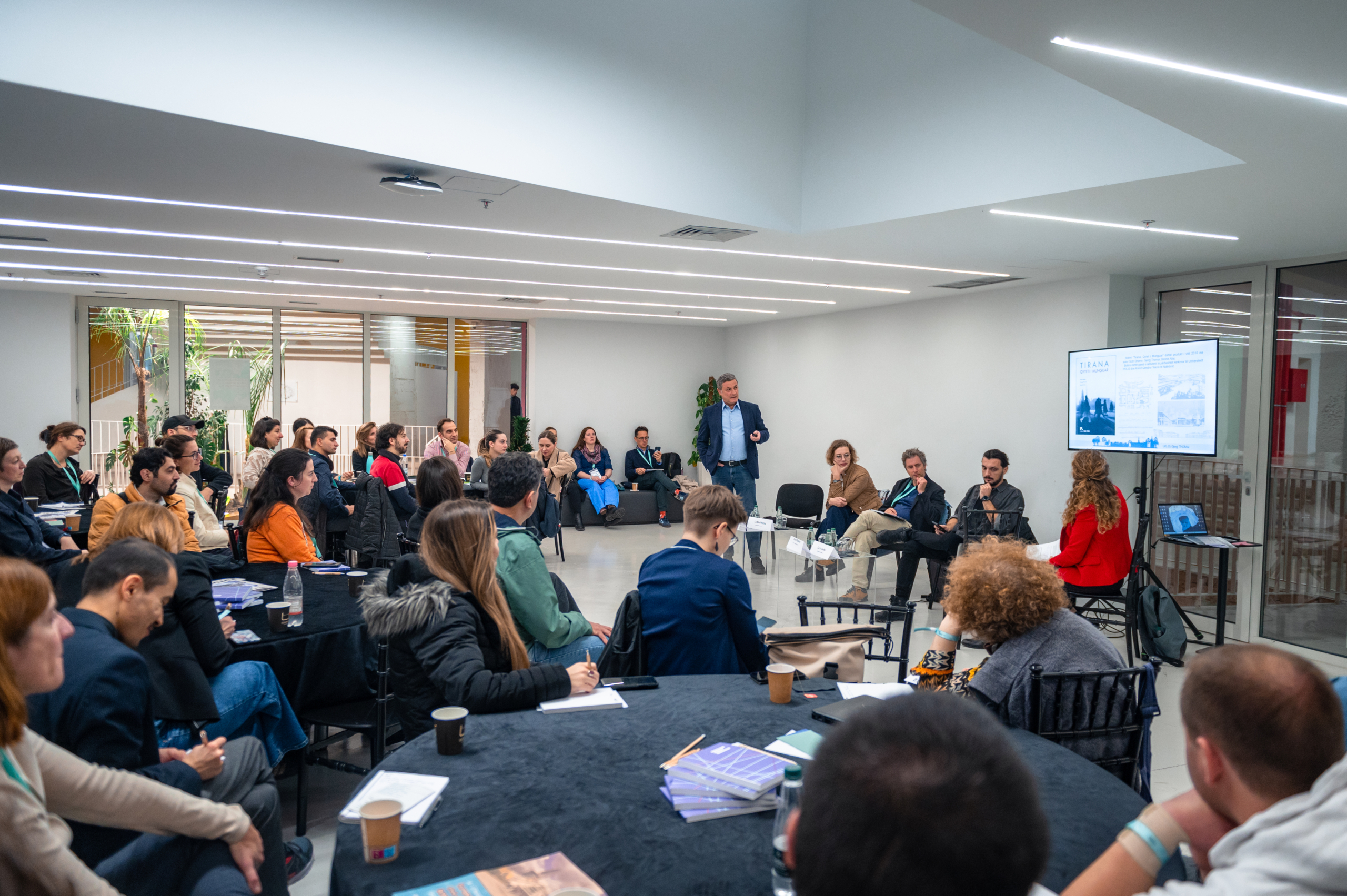 © THE CIVICS Innovation Hub | Geri Lula A diverse group of people attentively listens to a presentation during NECE Festival 2024. A speaker presents beside a screen displaying slides.