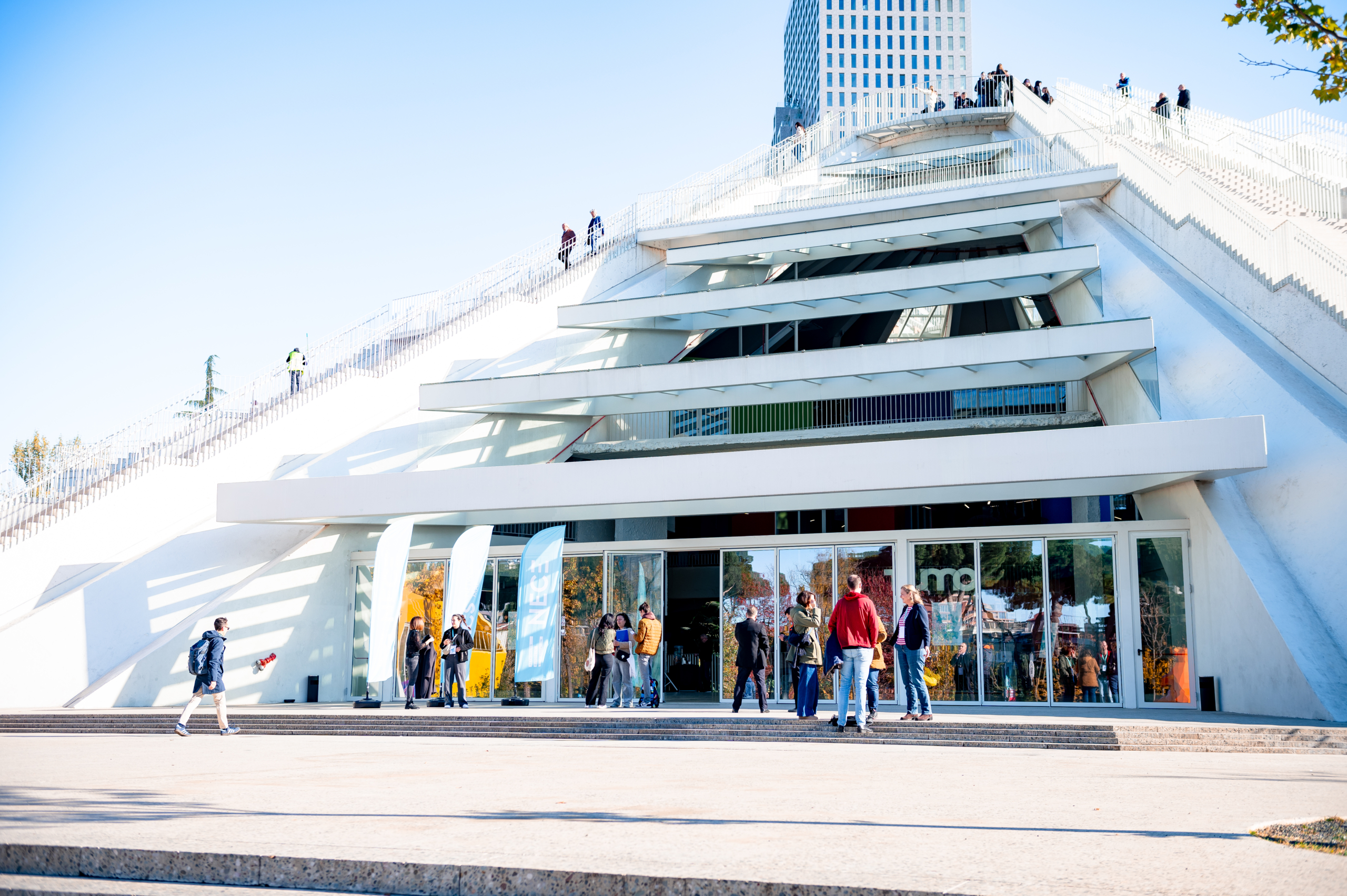 © THE CIVICS Innovation Hub | Geri Lula The Piramide building in Tirana - a modern building with angled architecture, bright white exterior under clear blue sky. People stroll, chat, creating a lively and sunny atmosphere.