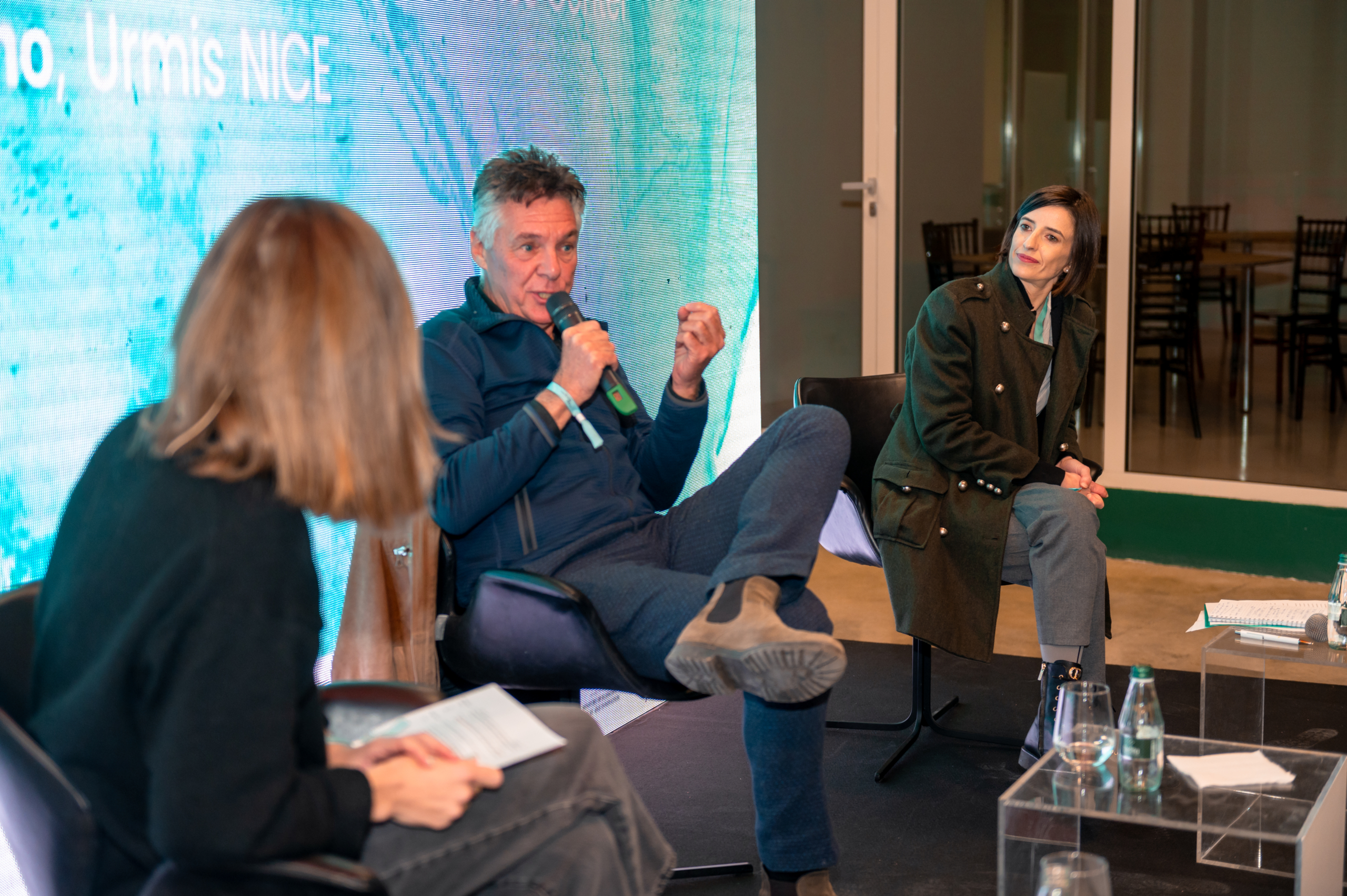 © THE CIVICS Innovation Hub | Geri Lula Three people are seated on a stage in a panel discussion during NECE Festival 2024. The man in the center speaks into a microphone, while the others listen attentively. There's a bright screen behind them and notes on a small table.