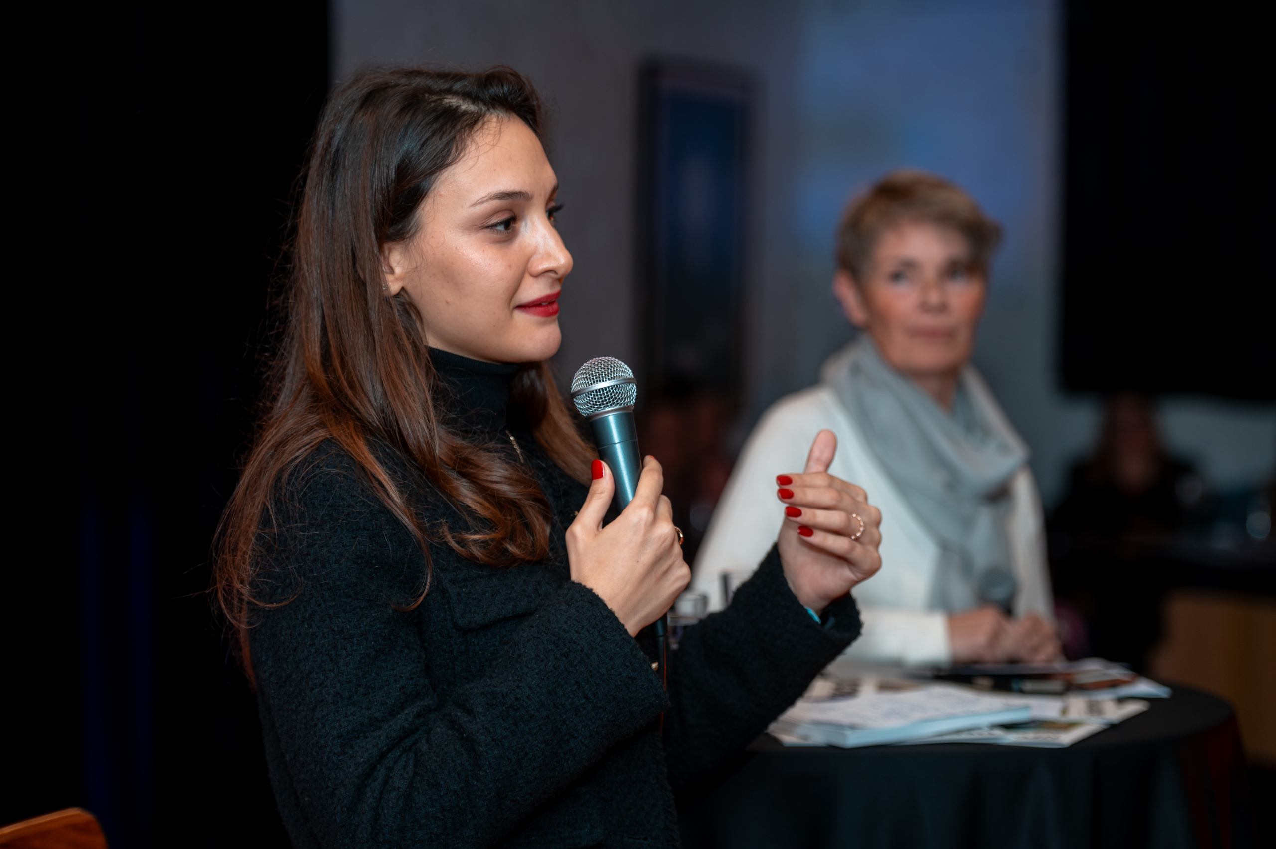 © THE CIVICS Innovation Hub | Geri Lula A woman in a black coat speaks into a microphone during NECE Festival 2024. A woman in the background listens intently.