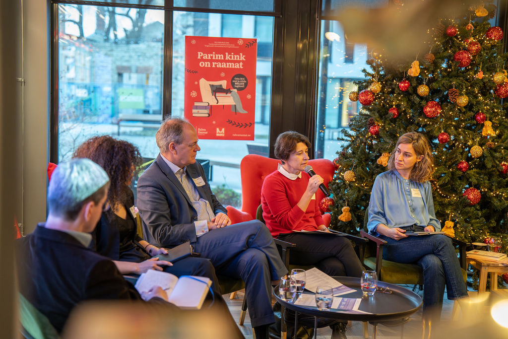 DSC03632-Enhanced-NR A group of five people engaged in a seated discussion during the NECE Lab in Tallinn, including one speaking into a microphone. They're in a cozy room with a Christmas tree adorned with red and gold ornaments.