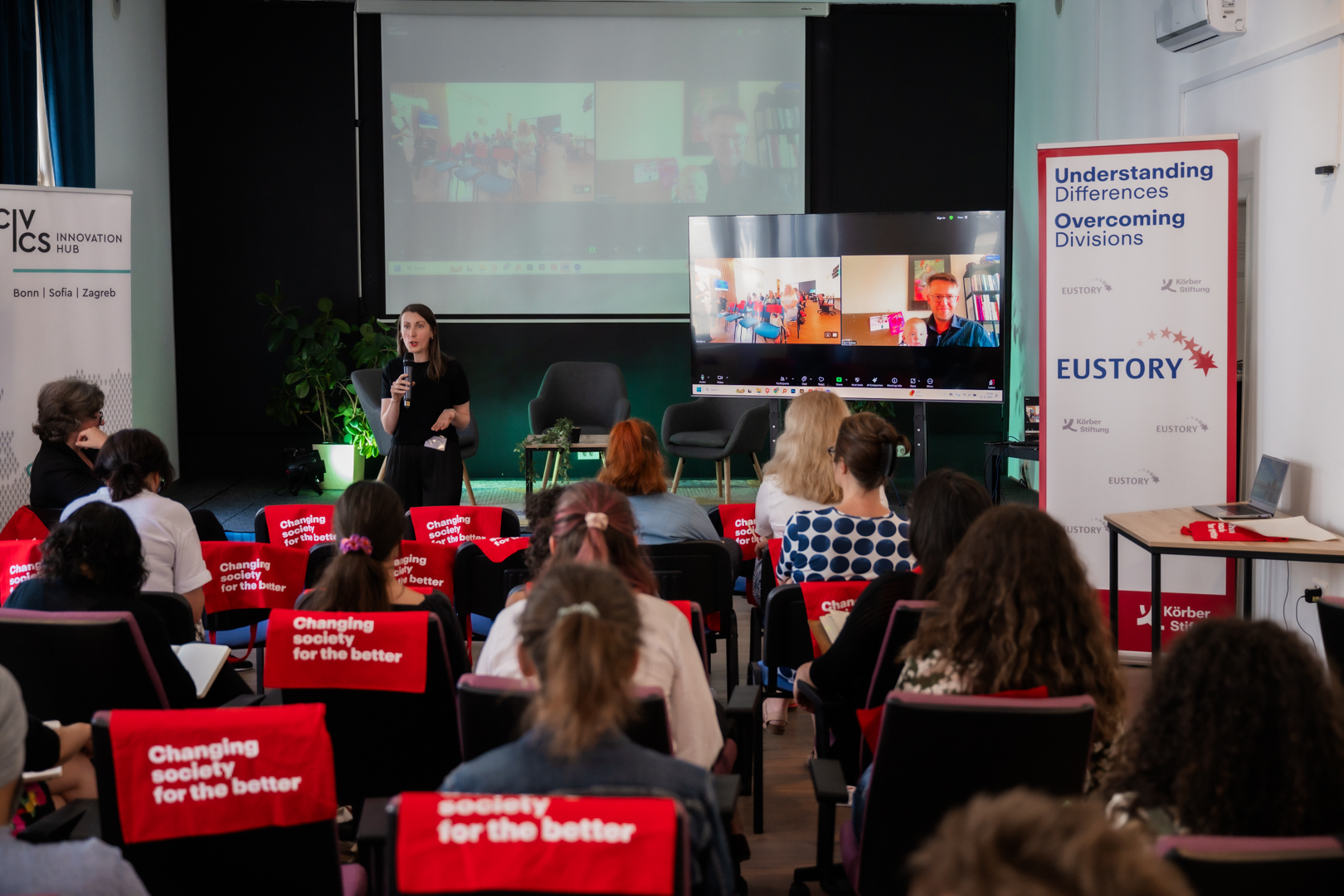 NECE Lab Sarajevo A woman speaks at a conference room with virtual attendees on a screen, surrounded by an audience seated in chairs with red covers. The atmosphere is engaged and focused.