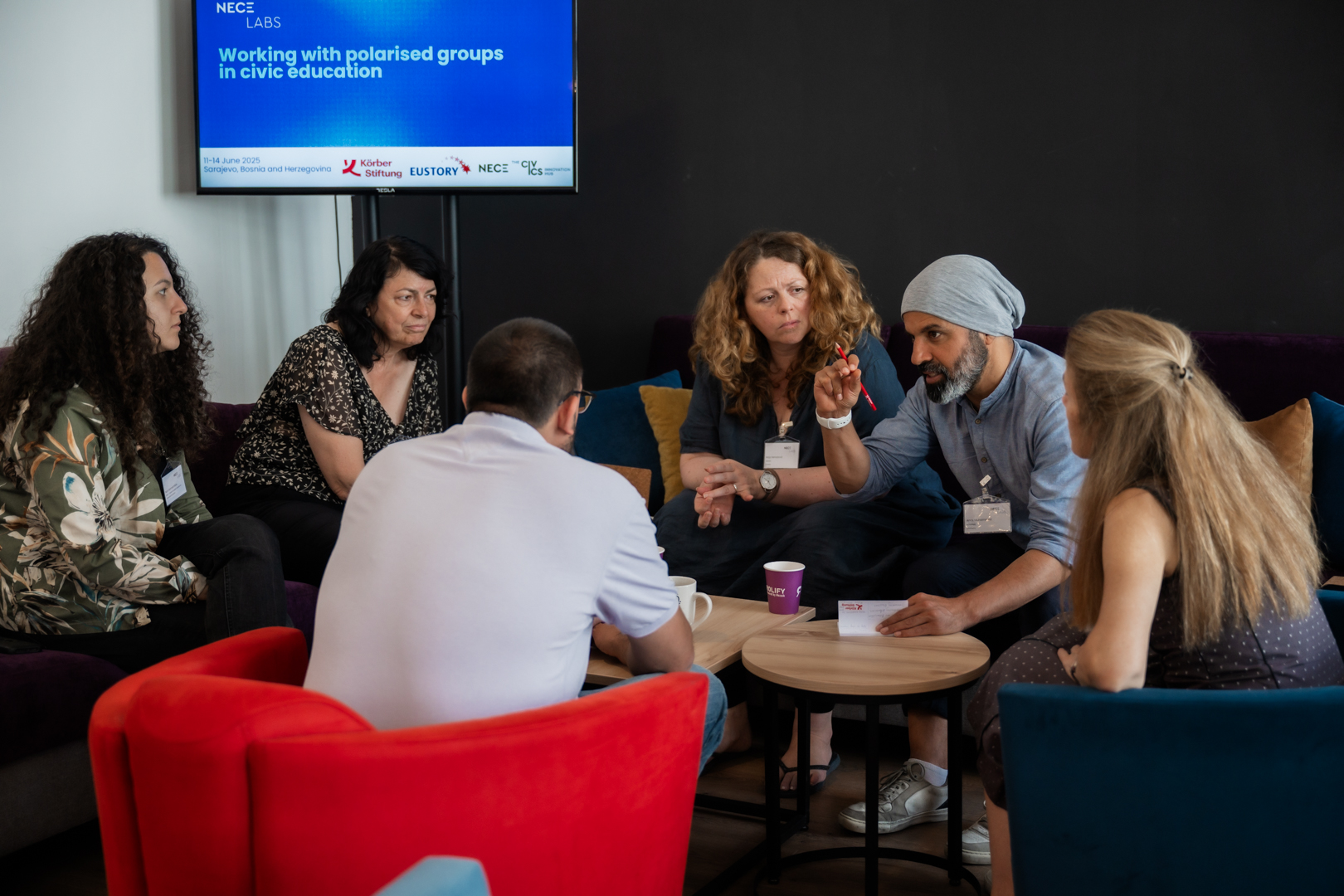 NECE Lab Sarajevo A diverse group of six people sits in a circle engaged in discussion. A screen in the background reads, "Working with polarised groups in civic education."