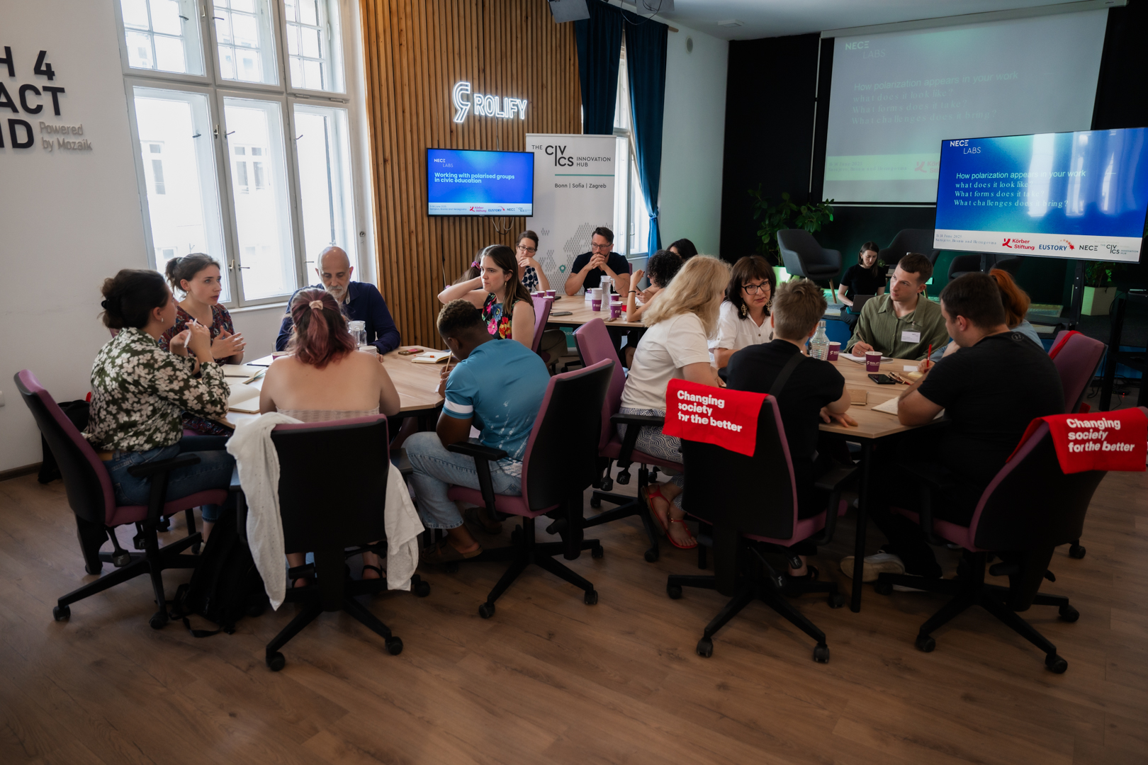 NECE Lab Sarajevo A group of diverse individuals sit at tables in a bright room, engaged in discussion. A screen displays text about working with polarized groups.