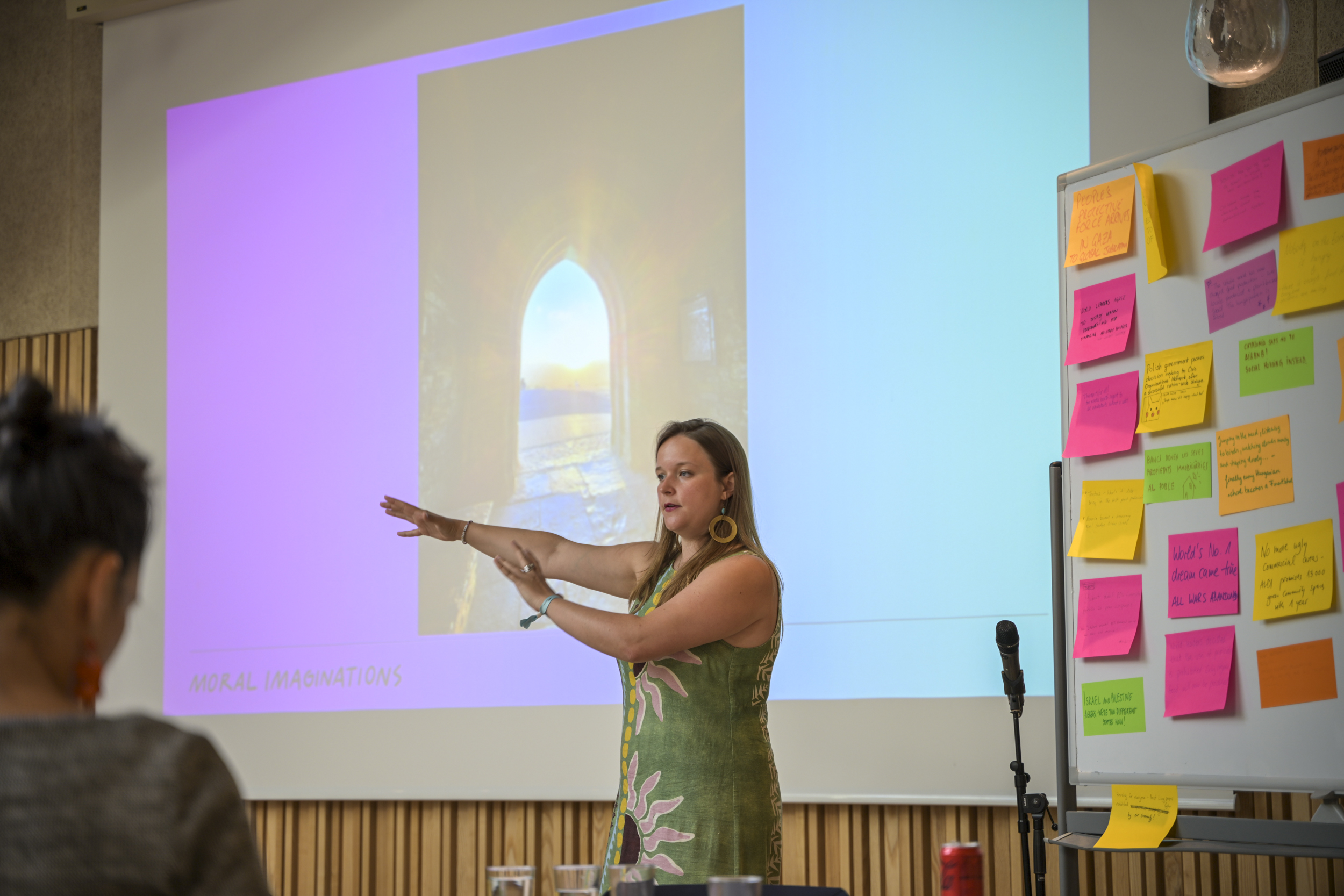 © THE CIVICS Innovation Hub / Werner Anderson Phoebe Tickell presents in a seminar room during a NECE Festival Masterclass. A board with colorful sticky notes is beside her.