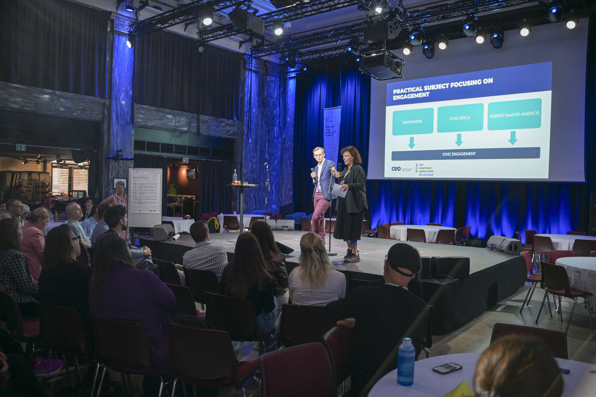 © THE CIVICS Innovation Hub / Werner Anderson A man and a woman are presenting on a stage during NECE Festival: HOPE. There is a large screen displaying engagement strategies. Audience members sit facing them in a dimly lit room.