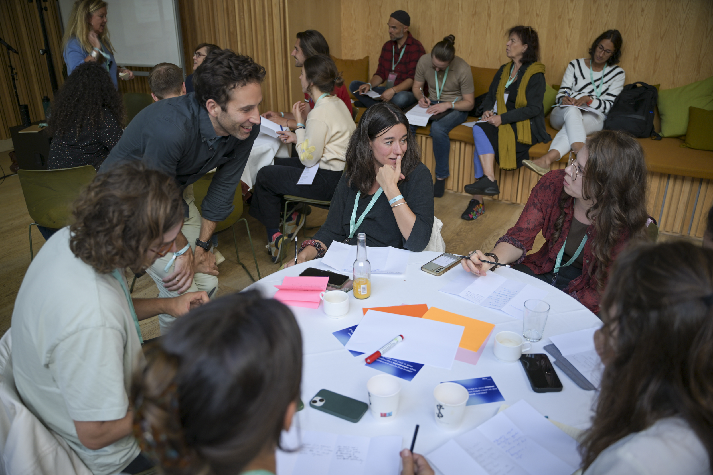 © THE CIVICS Innovation Hub / Werner Anderson A group of people gathers around a table, engaged in discussion with papers. Others are seated in the background, creating a collaborative and lively atmosphere.