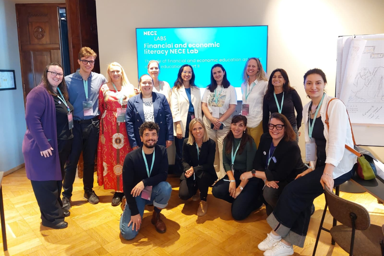 THE CIVICS Innovation Hub Group of 14 people pose indoors, smiling and wearing NECE Festival lanyards. Behind them, a screen displays "Financial and Economic Literacy NECE Lab."