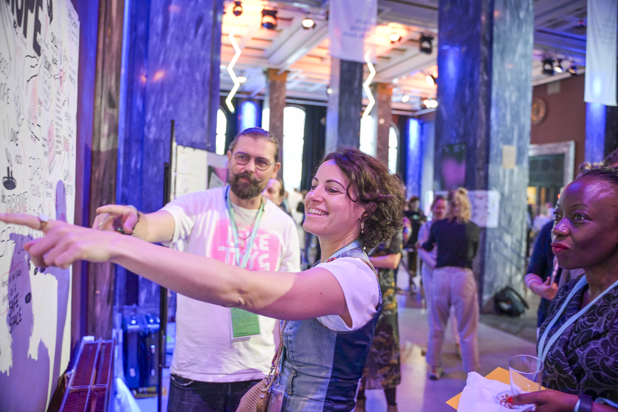 © THE CIVICS Innovation Hub / Werner Anderson Three individuals in a colorful, lively room engage with a large interactive board at the NECE Festival: HOPE. They appear engaged and joyful, pointing and discussing ideas.