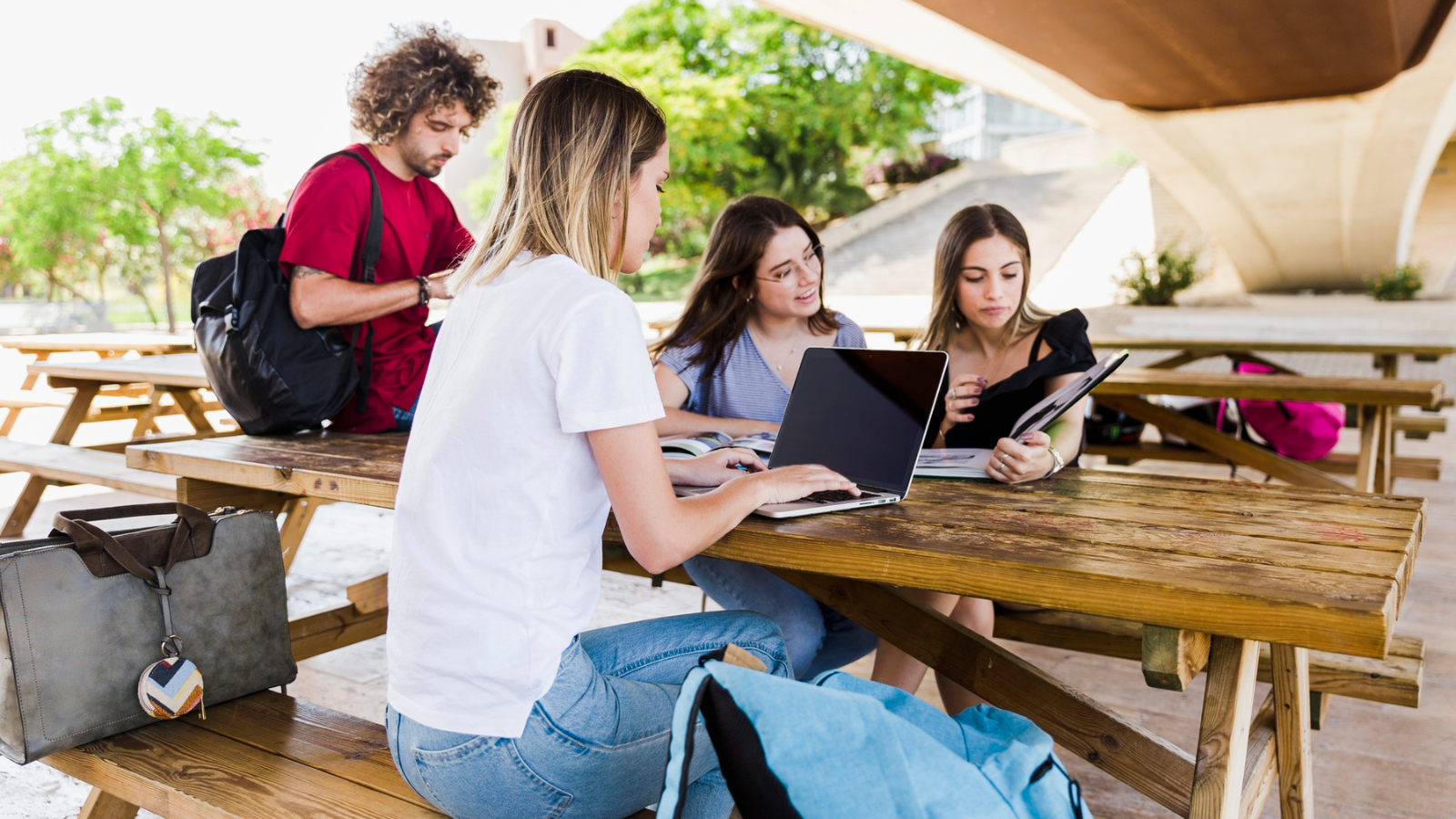 Four young adults study outdoors at a wooden picnic table under a bridge.