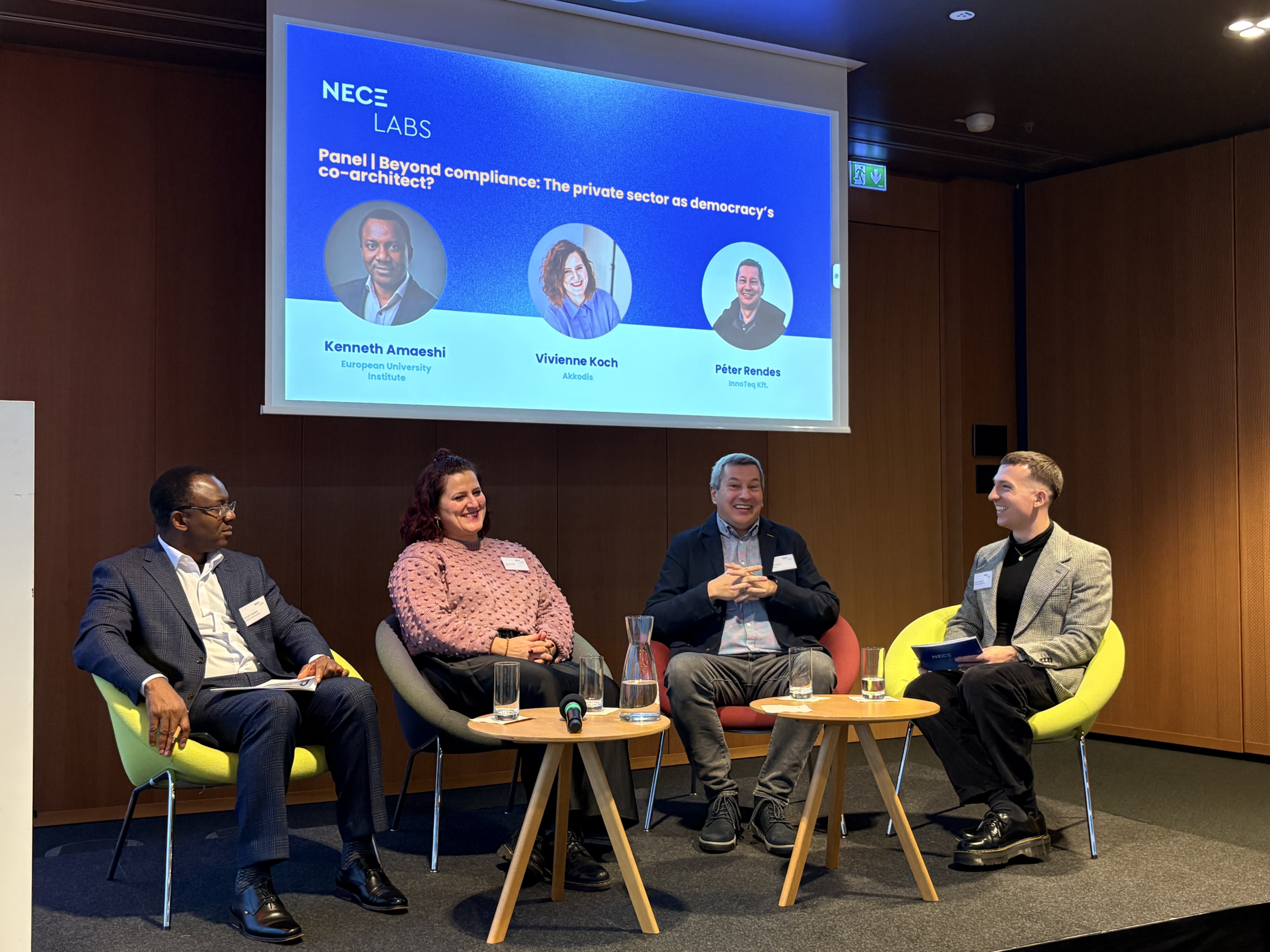 Four panelists sit on stage, smiling and engaged in discussion. Behind them, a screen displays the panel topic about the private sector and democracy.
