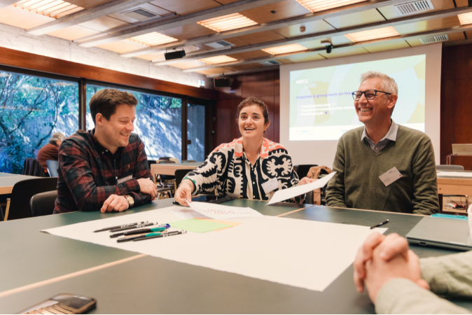 © THE CIVICS Innovation Hub / Tomás Nazaré Image showing three participants sitting on a table while doing group work. They are laughing, and there is some paper on the table.